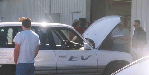 SUV with "EV" on the passenger door. The hood is open and two people are looking under it.