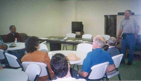 Several people sitting in white, folding chairs at multiple tables. One person is standing as most seated look toward him.
