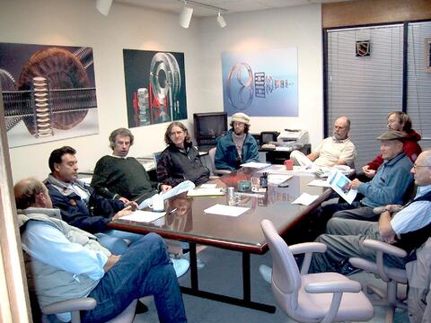 People seated around a long glass-topped table in a conference room filled with mechanical/technology art.